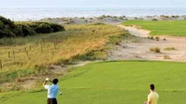 Two people are playing golf on a grassy course near the coast, with one person swinging a club while the other watches.