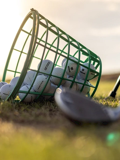 A green basket of golf balls lies on the grass with a golf club head in the foreground.