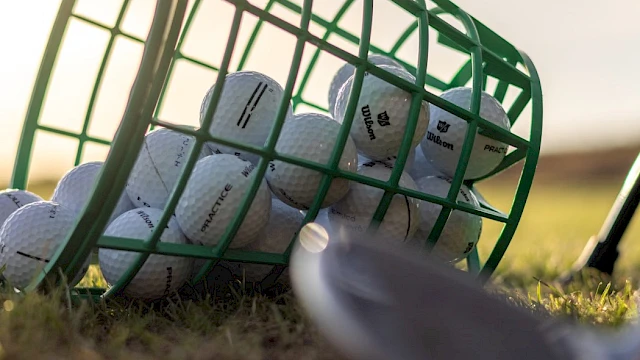 A green basket of golf balls lies on the grass with a golf club head in the foreground.