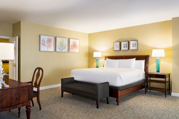 The image shows a cozy bedroom with a large bed, wooden furniture, lamps, and artwork on the walls, creating a warm and inviting atmosphere.