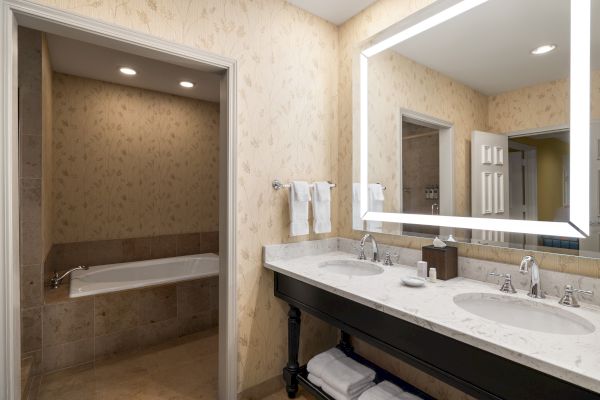 A bathroom with a double sink, illuminated mirror, towels, and a bathtub in an adjacent alcove, featuring floral wallpaper and tile accents.