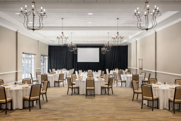 A banquet hall with round tables set for a formal event, chandeliers on the ceiling, and a large projector screen at the front.
