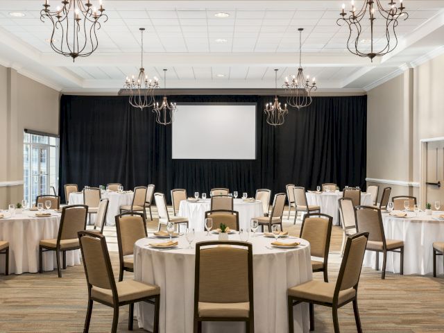 The image shows an elegantly set banquet hall with round tables, chairs, chandeliers, and a large screen at the front.