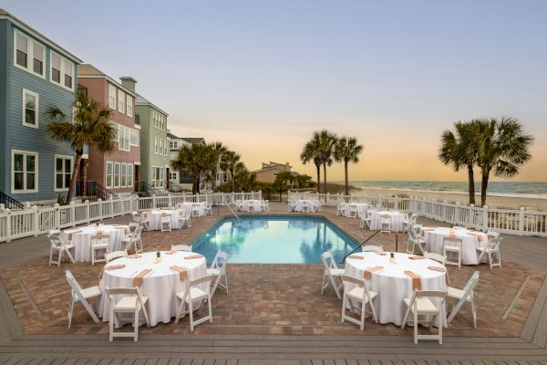 A beachside setup with round tables covered in white cloth surrounding a pool, palm trees, colorful houses, and ocean view at sunset.