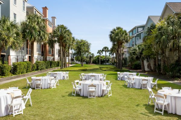 Outdoor event setup with round tables and white chairs on grass, surrounded by palm trees and buildings under a clear blue sky.