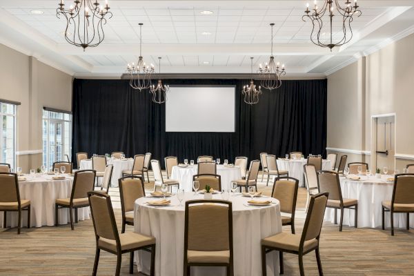 A banquet hall with round tables set for an event, featuring chandeliers, a projector screen, and neutral decor.