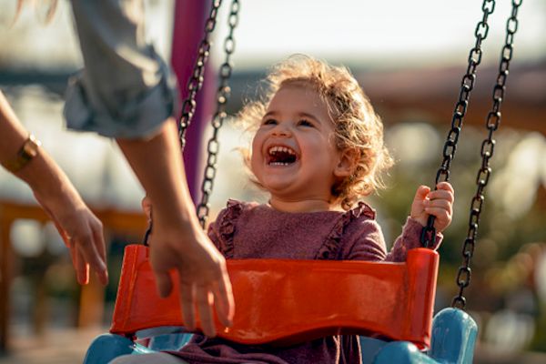 A child is joyfully swinging and smiling while someone pushes them at a playground.