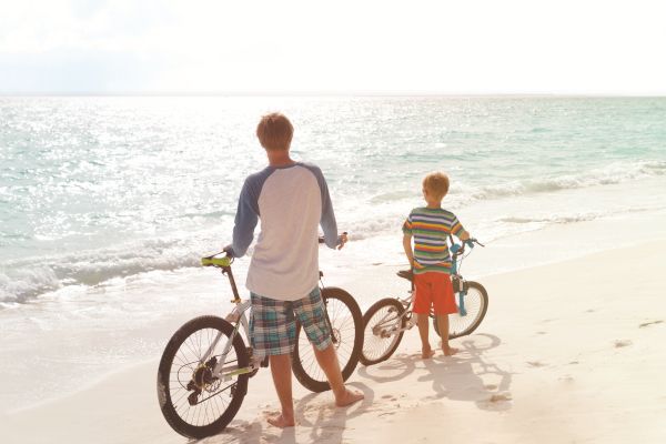 Two people with bikes on a sunny beach, facing the ocean.