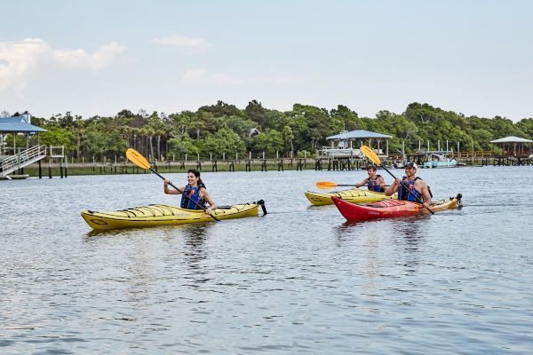 Three people kayaking on a calm body of water, with trees and docks in the background, enjoying an outdoor activity on a sunny day.