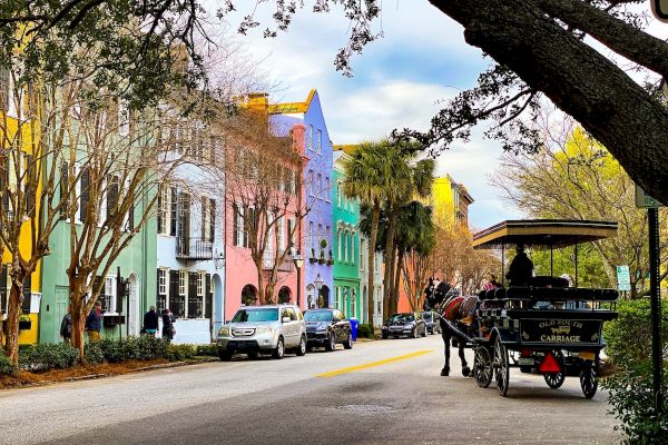 A street with colorful historic buildings, parked cars, and a horse-drawn carriage under trees, creating a picturesque scene.