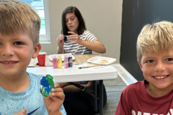 Children are showing painted crafts while sitting at a table with art supplies in a room.