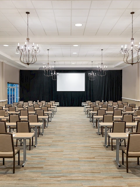 A conference room with rows of tables and chairs facing a screen, under chandeliers, ready for a meeting or presentation.