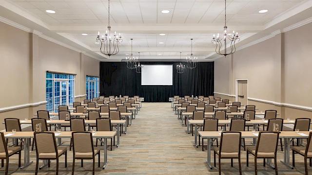A conference room with rows of tables and chairs facing a screen, under chandeliers, ready for a meeting or presentation.
