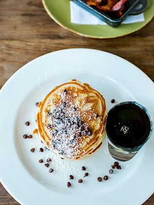 A stack of pancakes with chocolate chips and a dusting of powdered sugar, served with syrup, and a side of bacon nearby.